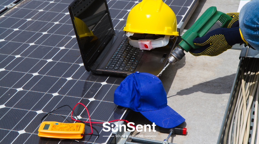 Solar panel installation tools including a laptop, multimeter, hard hat, drill, and work gloves laid out on a solar panel.